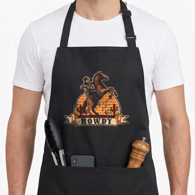 Tablier de cuisine noir avec illustration d'un cowboy chevauchant un cheval cabré devant des cactus et le texte Howdy.
