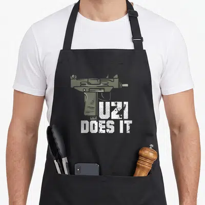 Tablier de cuisine noir avec illustration pistolet Uzi et texte Uzi Does It en blanc texturé.