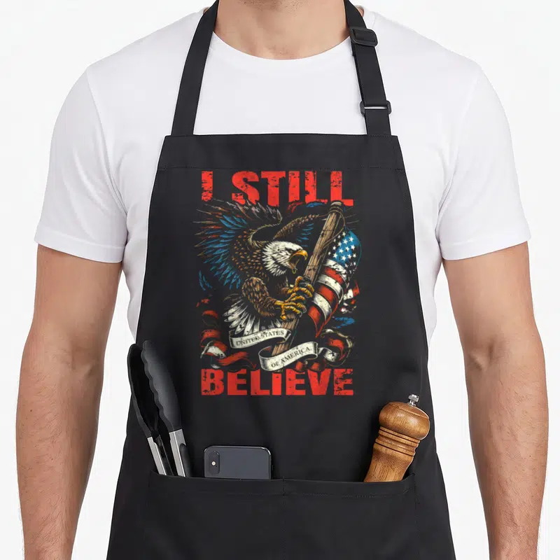 Tablier de cuisine noir avec aigle tenant le drapeau américain et texte rouge I Still Believe.