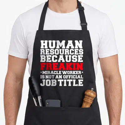 Tablier de cuisine noir avec le texte blanc et rouge Human Resources Because Freakin Miracle Worker Is Not An Official Job Title imprimé au centre.