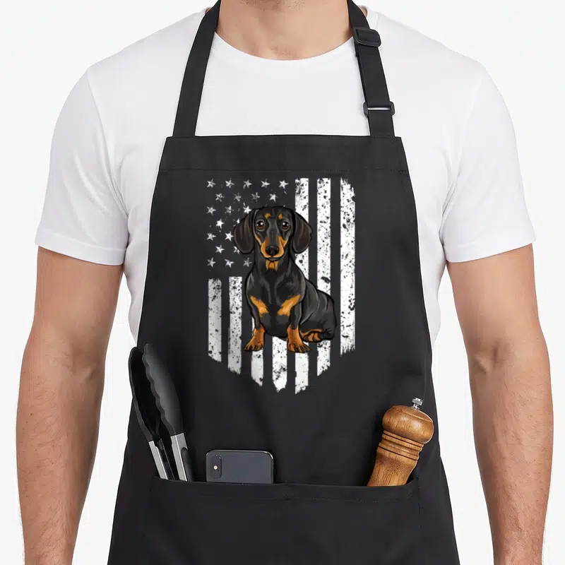 Tablier de cuisine noir avec illustration d'un teckel noir devant un drapeau américain stylisé en blanc et gris.