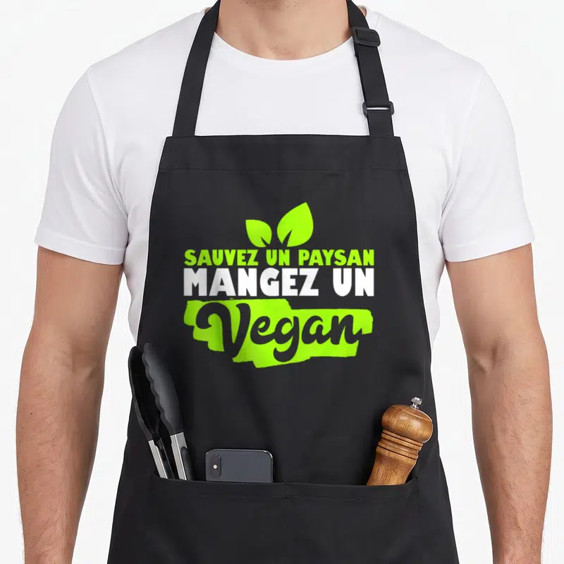 Tablier de cuisine noir avec l'inscription verte et blanche « Sauvez un paysan mangez un vegan ». Illustration de deux feuilles au-dessus du texte.