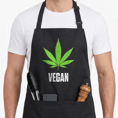 Tablier de cuisine noir orné d'une feuille de cannabis verte et du texte VEGAN en blanc au bas du motif central.