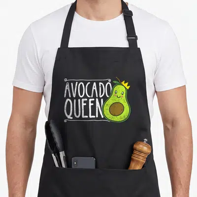 Tablier de cuisine noir avec illustration d'un avocat souriant portant une couronne jaune et le texte Avocado Queen en lettres blanches.
