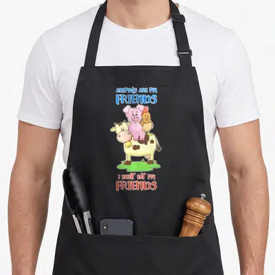 Tablier de cuisine noir avec une vache, un cochon rose et une poule illustrés en couleur avec du texte bleu et rouge.
