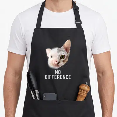 Tablier de cuisine noir avec un visage divisé entre un porcelet rose et un chat tigré au-dessus du texte No Difference en blanc.