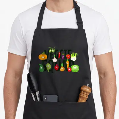 Tablier de cuisine noir avec linscription Veggie Squad et des légumes dessinés avec des visages souriants colorés.