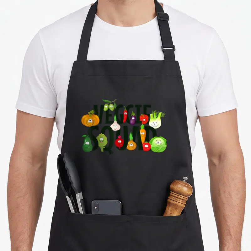 Tablier de cuisine noir avec linscription Veggie Squad et des légumes dessinés avec des visages souriants colorés.