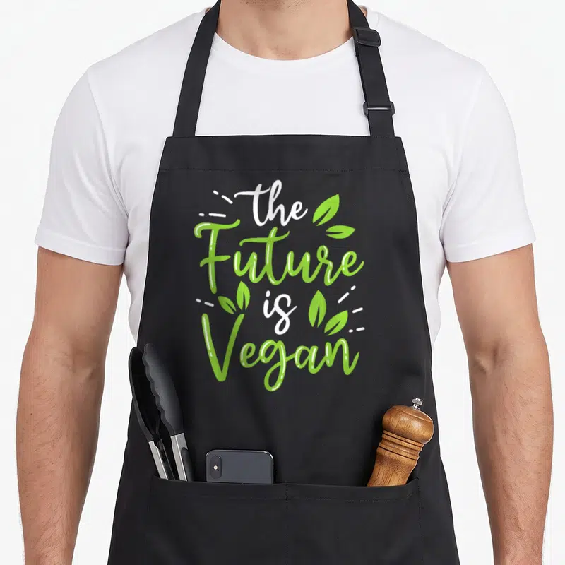 Tablier de cuisine noir avec le texte The Future Is Vegan en vert et blanc entouré de feuilles stylisées.