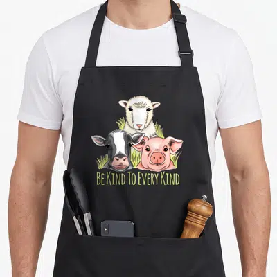 Tablier de cuisine noir avec illustration de mouton, vache et cochon sous le texte Be Kind To Every Kind en jaune.