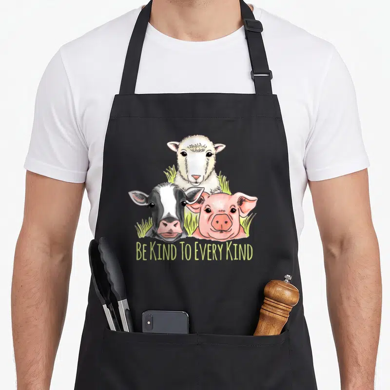 Tablier de cuisine noir avec illustration de mouton, vache et cochon sous le texte Be Kind To Every Kind en jaune.