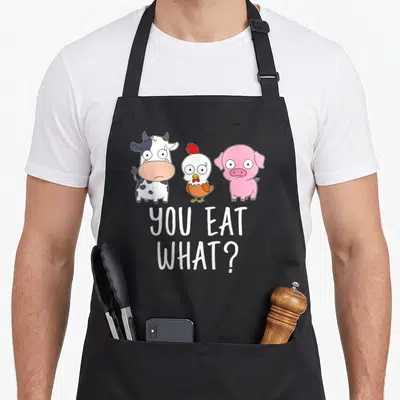 Tablier de cuisine noir avec une vache, un poulet et un porc dessinés en cartoon sous le texte You Eat What en lettres blanches.
