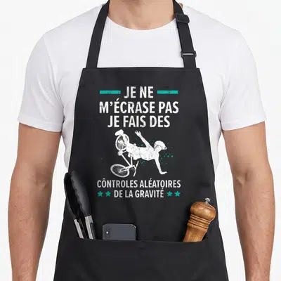 Tablier de cuisine noir avec illustration blanche d'un cycliste qui tombe et texte humoristique sur fond gris uni.