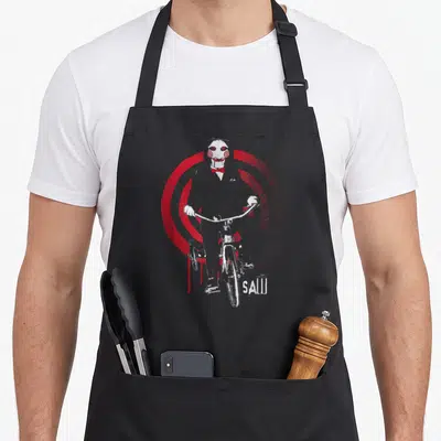 Tablier de cuisine noir avec illustration du personnage Saw Jigsaw montant un vélo devant des cercles rouges concentriques.