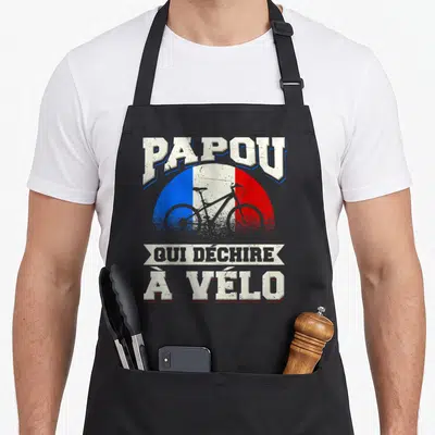 Tablier de cuisine noir avec motif Papou qui déchire à vélo sur fond tricolore et silhouette de bicyclette grise.
