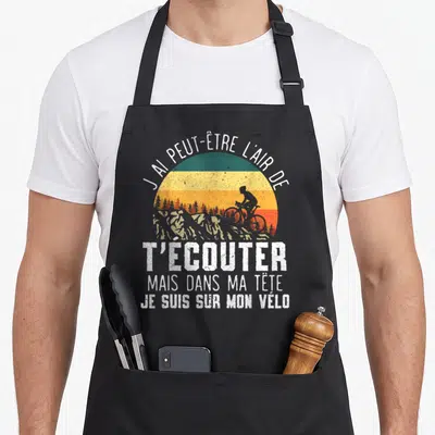 Tablier de cuisine noir avec illustration vintage d'un cycliste et texte blanc humoristique sur fond de soleil dégradé.