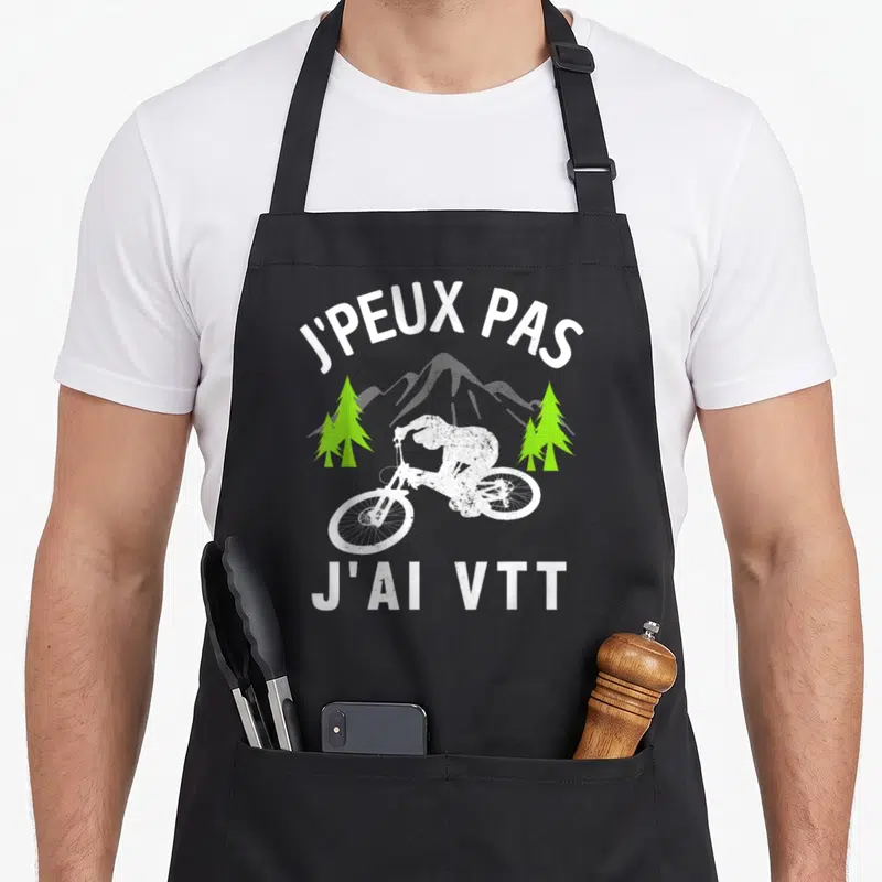 Tablier de cuisine noir avec cycliste VTT blanc, arbres verts et texte J'peux pas j'ai VTT en typographie blanche.