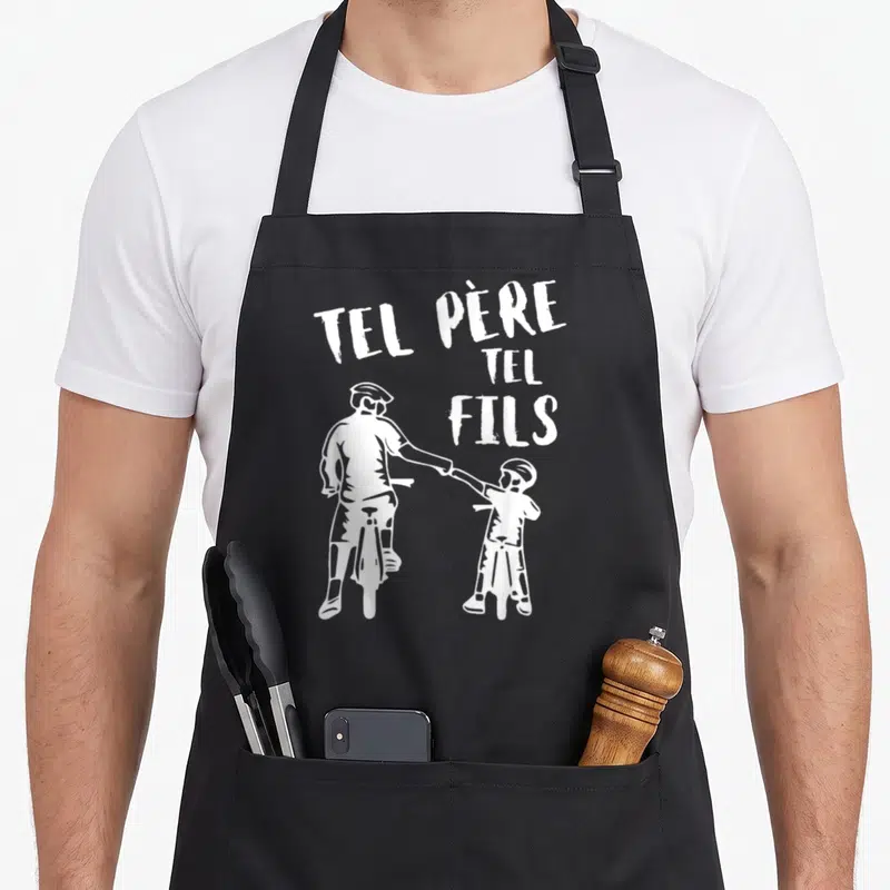 Tablier de cuisine noir avec illustration blanche d'un père et un fils à vélo se tenant la main sous le texte Tel Père Tel Fils.