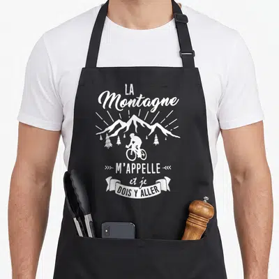 Tablier de cuisine noir avec illustration blanche d'un cycliste devant des montagnes et le texte La Montagne m'appelle et je dois y aller.