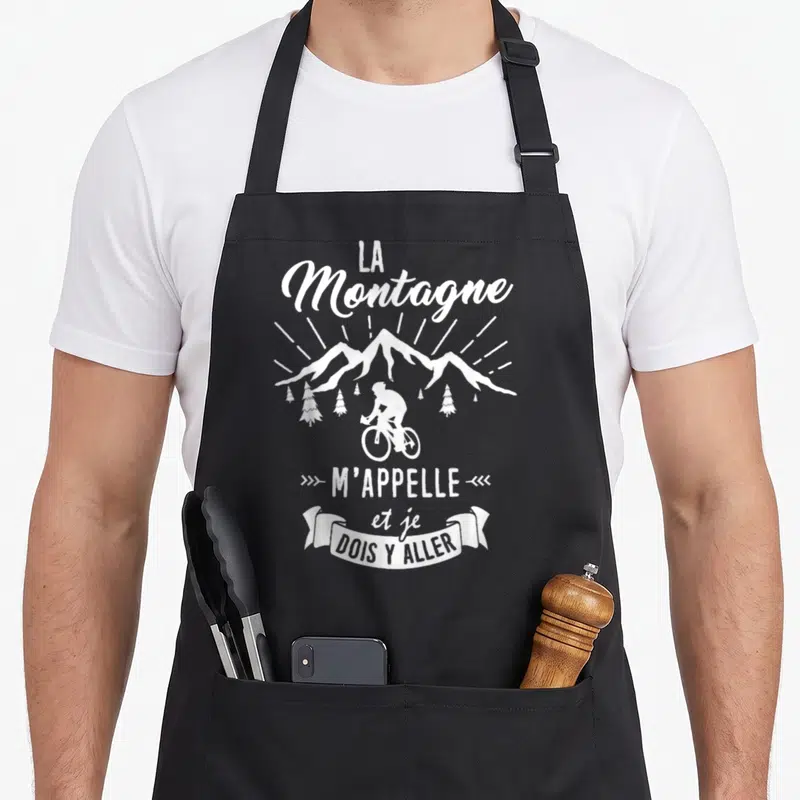 Tablier de cuisine noir avec illustration blanche d'un cycliste devant des montagnes et le texte La Montagne m'appelle et je dois y aller.