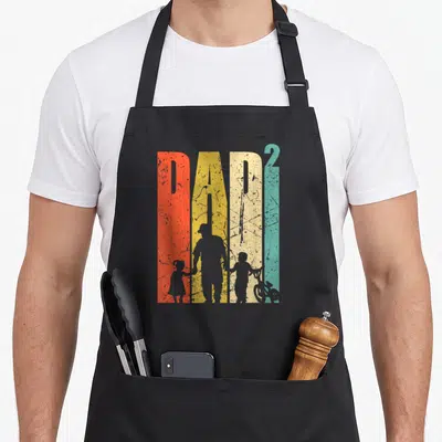 Tablier de cuisine noir avec illustration Dad 2 montrant un père tenant la main d'un enfant près d'un vélo BMX.