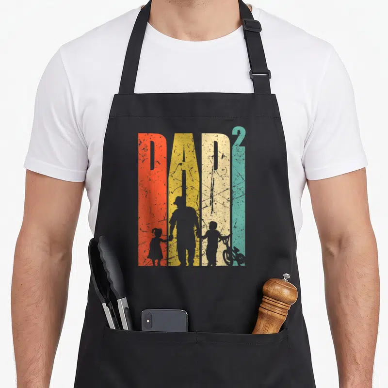 Tablier de cuisine noir avec illustration Dad 2 montrant un père tenant la main d'un enfant près d'un vélo BMX.