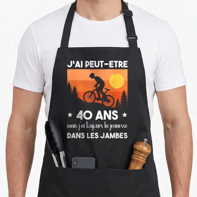Tablier de cuisine noir avec silhouette de cycliste, soleil orange et texte blanc célébrant les 40 ans avec humour.