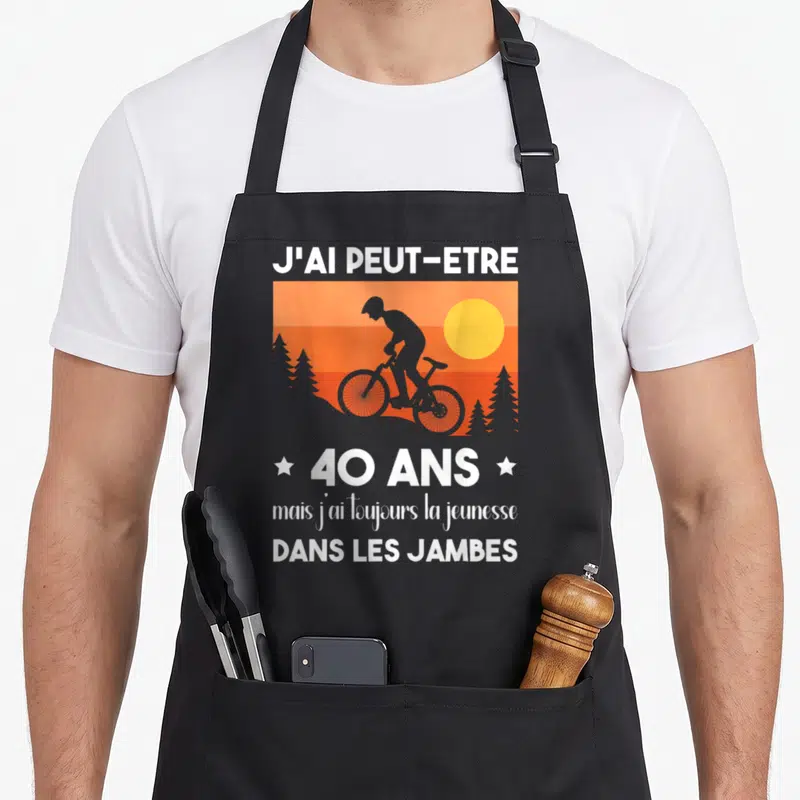 Tablier de cuisine noir avec silhouette de cycliste, soleil orange et texte blanc célébrant les 40 ans avec humour.