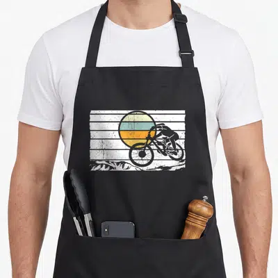 Tablier de cuisine noir avec illustration d'un cycliste MTB en descente devant un soleil coloré.