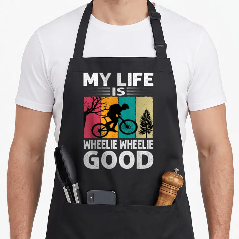 Tablier de cuisine noir avec silhouette cycliste sur fond coloré et texte blanc My Life Is Wheelie Wheelie Good.