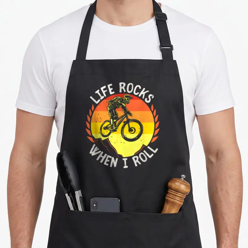 Tablier de cuisine noir avec illustration cycliste VTT et texte Life Rocks When I Roll sur fond de soleil orange jaune.