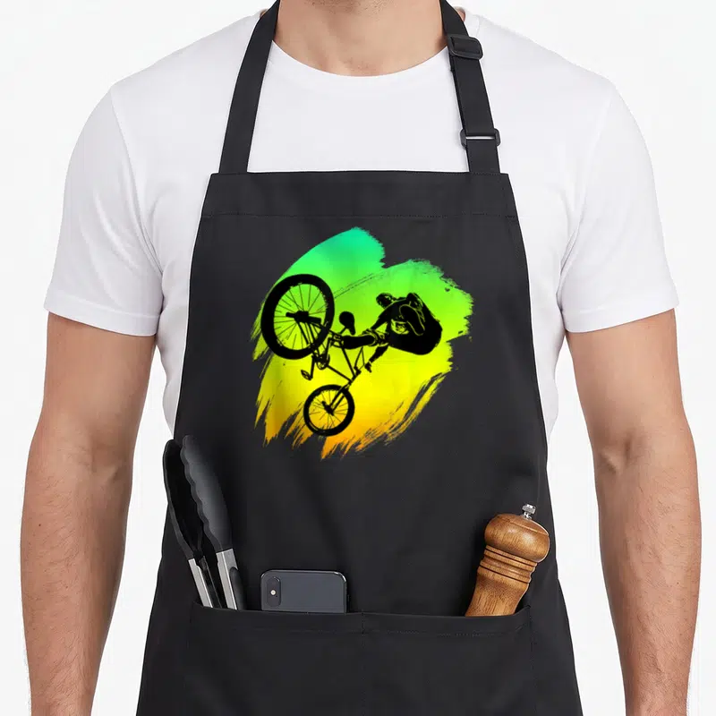 Tablier de cuisine noir avec silhouette cycliste effectuant un saut acrobatique sur fond dégradé vert et jaune.