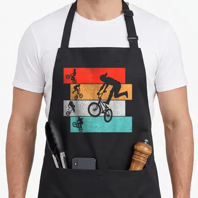 Tablier de cuisine noir avec silhouettes de cyclistes BMX sur bandes rouges, oranges, blanches et turquoise texturées.