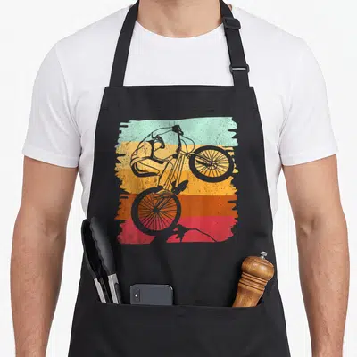 Tablier de cuisine noir avec illustration cycliste VTT effectuant un wheelie sur fond de bandes colorées délavées.