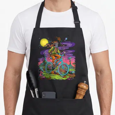 Tablier de cuisine noir avec illustration colorée d'un personnage en cyclisme traversant un décor psychédlique aux teintes violettes et vertes.