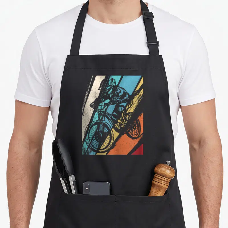 Tablier de cuisine noir avec illustration d'un cycliste VTT en action sur fond de bandes diagonales colorées.