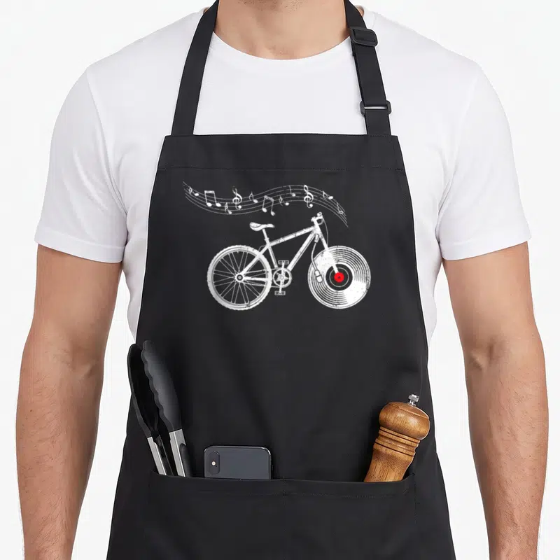 Tablier de cuisine noir avec illustration blanche d'un vélo BMX dont la roue avant est un disque vinyle rouge et des notes de musique.
