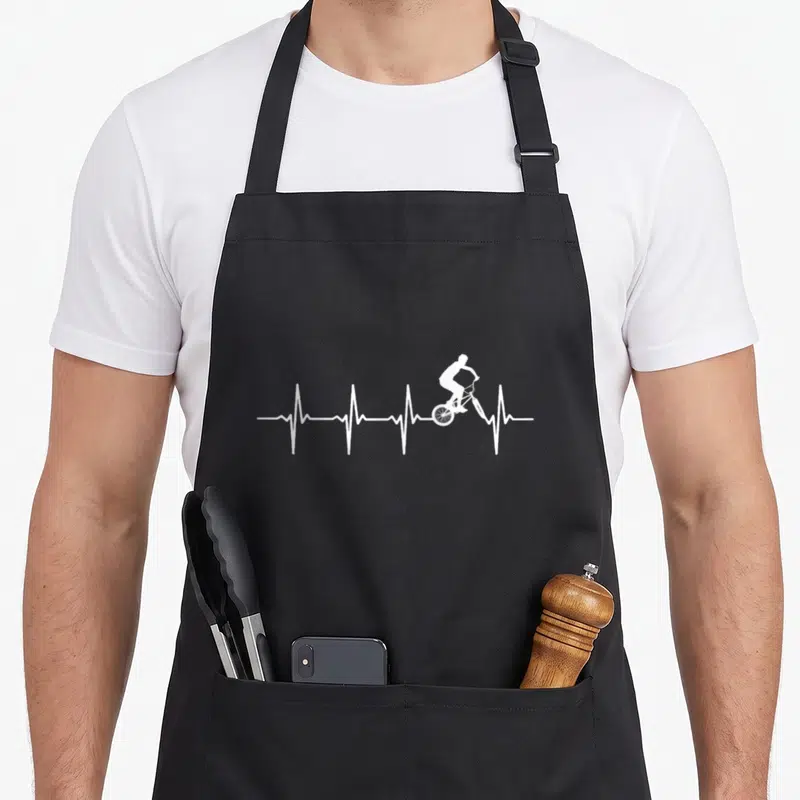 Tablier de cuisine noir avec un graphisme blanc représentant un bicrosseur sur une ligne de battement de cœur.