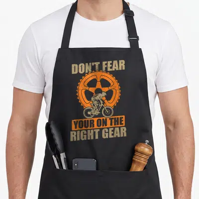 Tablier de cuisine noir avec illustration d'un cycliste sur une grande pignon orange et texte beige.