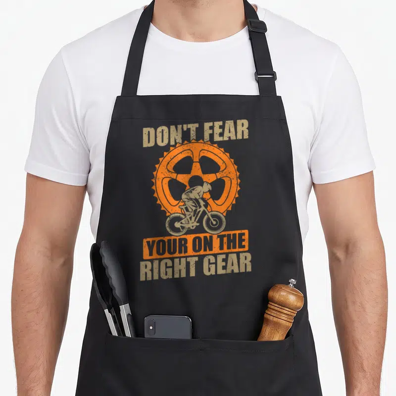 Tablier de cuisine noir avec illustration d'un cycliste sur une grande pignon orange et texte beige.