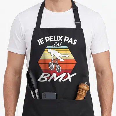Tablier de cuisine noir avec illustration d'un cycliste BMX blanc sur fond hexagonal aux couleurs dégradées orange et jaune.