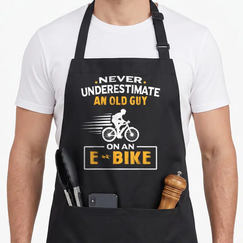 Tablier de cuisine noir avec silhouette cycliste et texte blanc et orange Never Underestimate An Old Guy On An E Bike