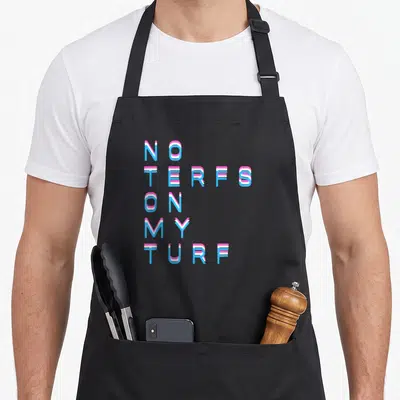 Tablier de cuisine noir avec le texte No Terfs On My Turf en lettres bleues et roses stylisées.