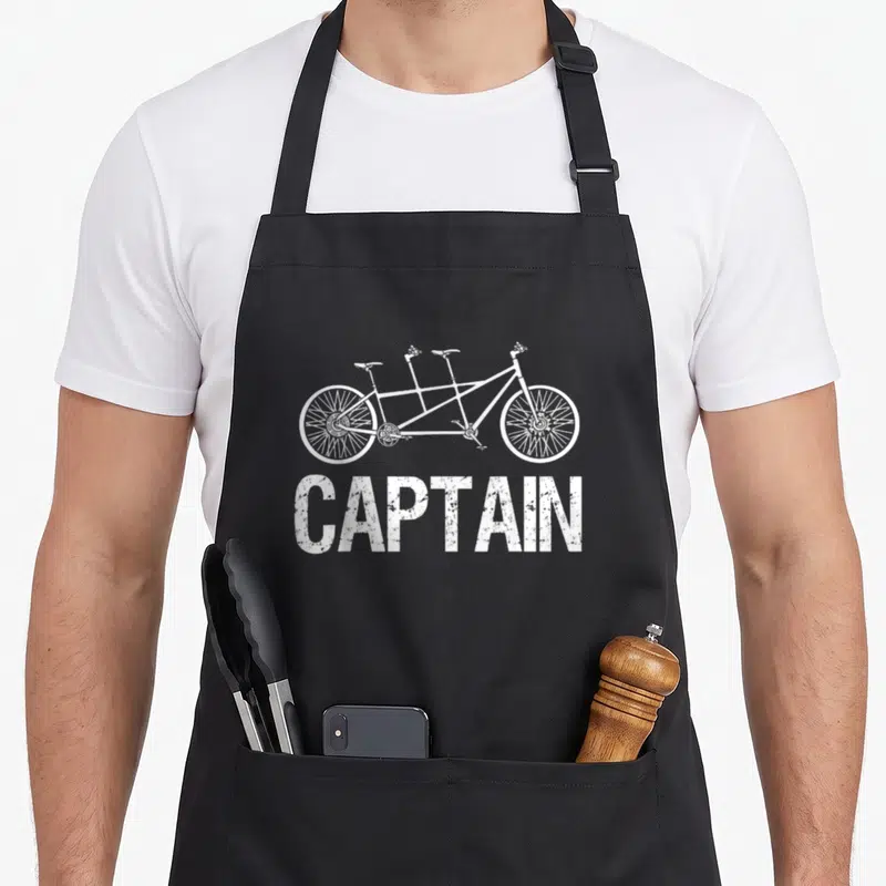 Tablier de cuisine noir avec illustration blanche d'un vélo tandem et l'inscription CAPTAIN en lettres capitales texturées.