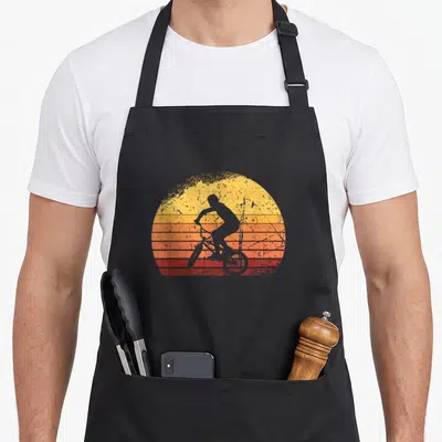 Tablier de cuisine noir avec illustration d'un cycliste BMX en silhouette sur un soleil couchant dégradé orange et rouge, style vintage