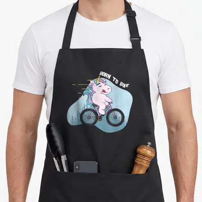 Tablier de cuisine noir avec une licorne cycliste rose et l'inscription Born to Bike sur fond bleu clair.