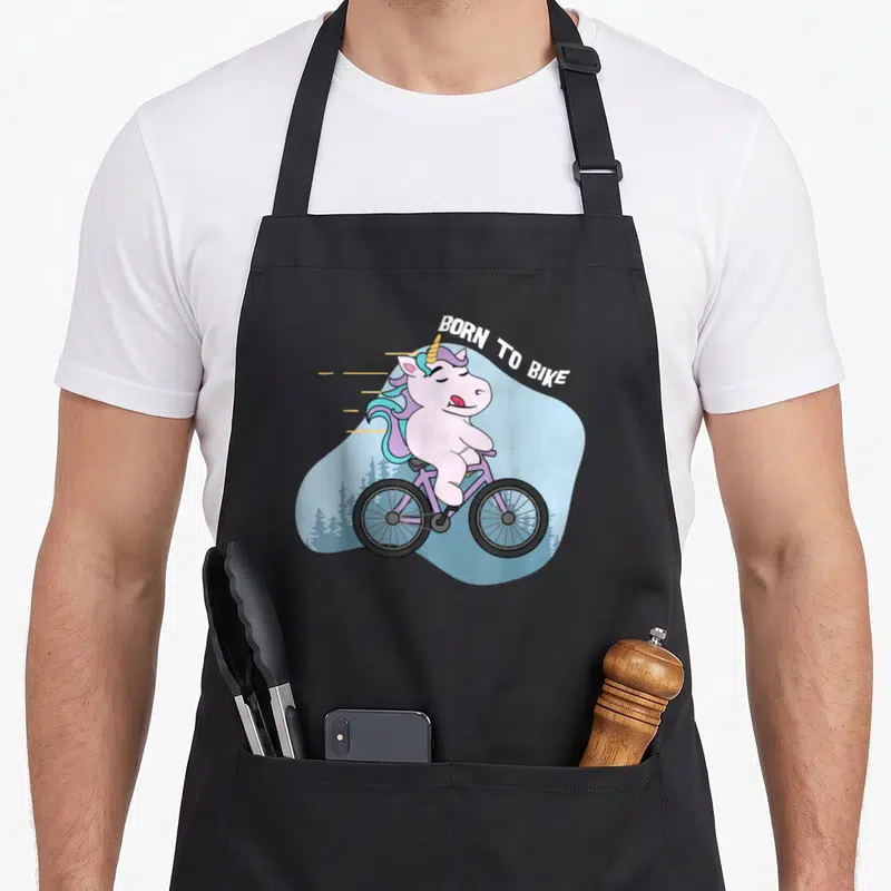 Tablier de cuisine noir avec une licorne cycliste rose et l'inscription Born to Bike sur fond bleu clair.