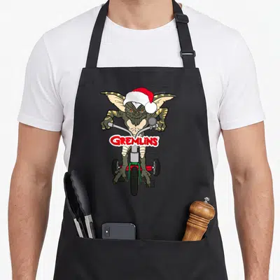 Tablier de cuisine noir avec un Gremlin en tricycle portant un bonnet de Noël et le logo GREMLINS.