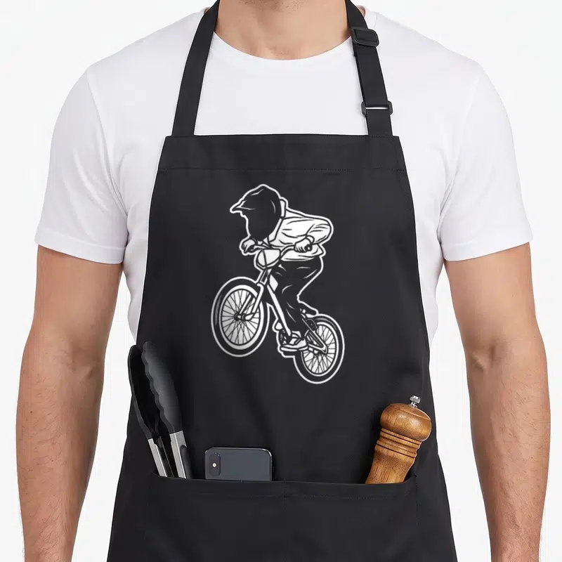 Tablier de cuisine noir avec illustration blanche d'un enfant en casque effectuant un wheelie sur un vélo BMX.