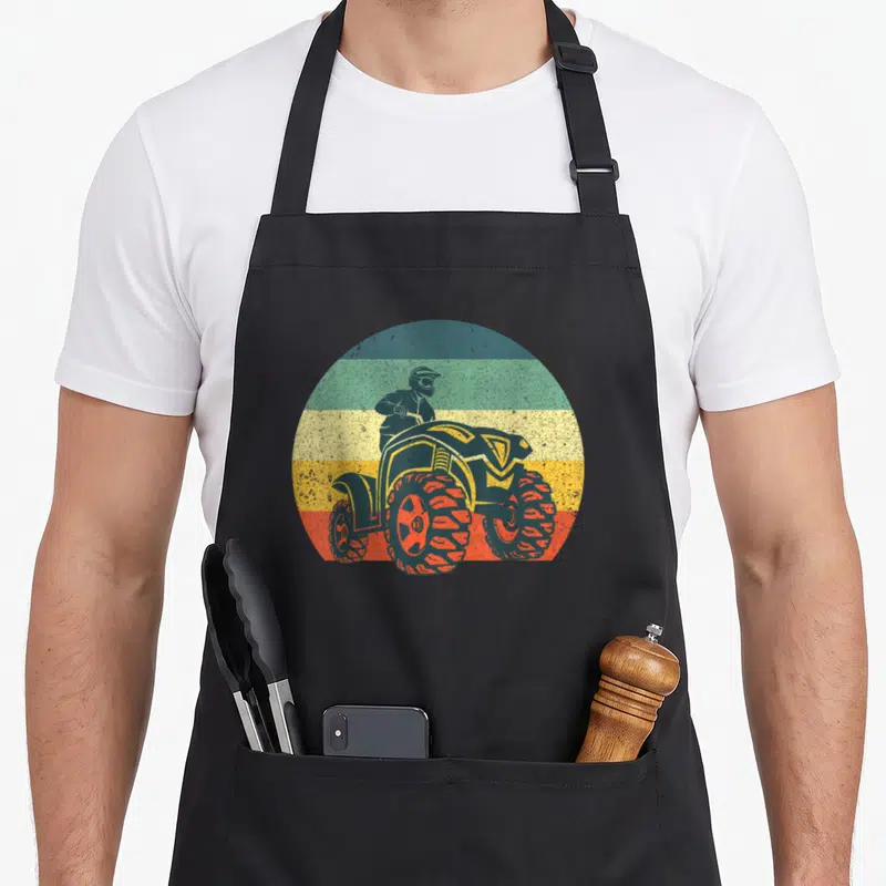 Tablier de cuisine noir avec illustration circulaire d'un quad ATV aux roues orange sur fond de bandes colorées vintage.